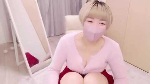 Media: Video of a short-haired Asian woman in a pink cardigan, wearing a face mask, sitting on a white rug in a minimalist room with light walls and a mirror.