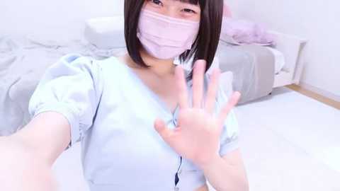 Media: Video of an East Asian woman with straight black hair, wearing a pink face mask, light blue shirt, and silver rings, holding her hand up in a bedroom with white walls, gray bed, and light wood floor.