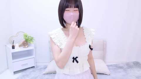 Media: Video of a young Asian woman with straight black hair, wearing a white frilled top with black bows, holding a pink mask to her face, in a minimalist, white-walled bedroom with a white nightstand, potted plant, and fluffy grey rug.