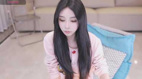 Media: Video of a young Asian woman with long black hair, wearing a pink sweater, kneeling on a white and blue checkered mat in a modern living room with beige couches.