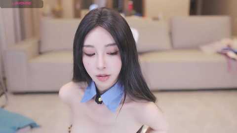 Media: Video of an Asian woman with long black hair, wearing a blue bow tie, sitting in a living room with beige furniture and a blue blanket.