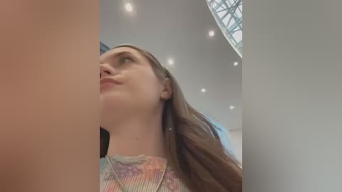 Video of a young woman with straight brown hair, wearing a colorful, textured top, standing in a modern, well-lit room with a circular skylight. The image is taken from a low angle, emphasizing her upward gaze.