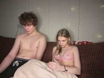 Video of a teenage boy with curly hair, shirtless, sitting next to a blonde girl in a pink bra on a brown couch.