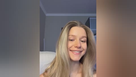 Media: Video of a smiling young woman with long, straight blonde hair, fair skin, and a small, subtle nose piercing. She wears a white top. Background shows a neutral-toned room with a white ceiling and a glimpse of a bed.