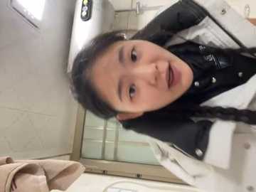 Media: Video of a young Asian woman with long black hair in braids, wearing a black and white uniform, looking directly at the camera in a bathroom with beige tiles and a beige towel.