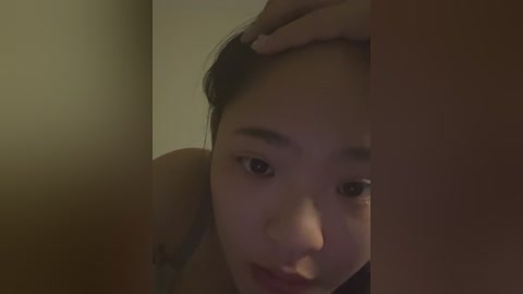 Media: Video of a young Asian woman with light skin, dark hair, and light makeup. She's resting her head on a brown wall, hand on her forehead, looking tired. Soft, dim lighting creates a warm, intimate atmosphere.