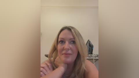 Media: Video of a middle-aged, blonde Caucasian woman with fair skin, holding her chin in a thoughtful pose, in a simple, beige room with a white ironing board and clothes in the background.