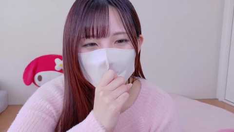 Media: Video of an East Asian woman with long, straight, brown hair, wearing a white surgical mask, pink sweater, and sitting in a room with a pink My Melody plush toy and white wall.