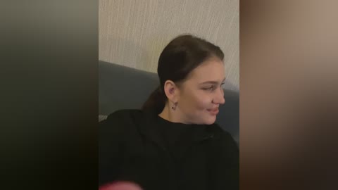 Media: Video of a young woman with light skin, brown hair tied back, wearing black clothing, smiling, sitting in a dimly lit room with textured beige walls and dark wooden furniture.