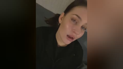 Media: Video of a Caucasian woman with fair skin and dark hair, wearing a black jacket, looking down with an expression of surprise or concern. The background is dark and blurred.