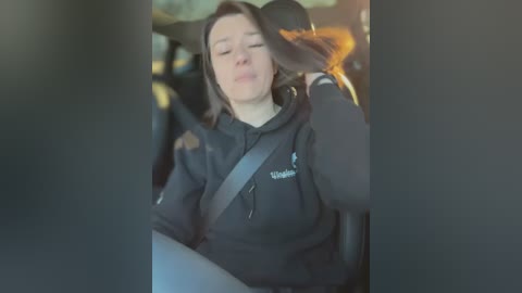 Media: Video of a woman with long brown hair, eyes closed, in a black hoodie, seated in a car, looking relaxed.