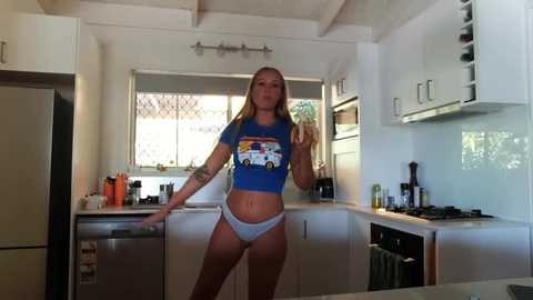 Media: Video of a fit, blonde woman with tattoos in a modern, white kitchen, wearing a blue crop top and grey panties, posing confidently.