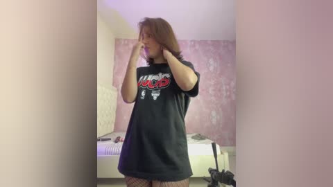 Video of a young woman with shoulder-length brown hair, wearing a black \"Rammstein\" t-shirt, fishnet stockings, and holding her hair in both hands. The background features pink floral wallpaper and a white bed.