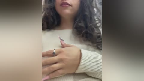 Media: Video of a light-skinned woman with curly dark hair, wearing a white sweater, holding her chest. Her left hand, adorned with a ring, rests on her heart. The background is blurred.