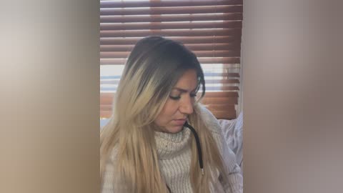 Media: Video of a Latina woman with long, straight, blonde hair, wearing a beige knit sweater, looking down, in a dimly lit room with wooden blinds.