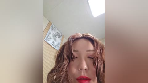 Media: A video of an Asian woman with wet, shoulder-length auburn hair, wearing bright red lipstick, captured from a low angle. The background features a beige wall with a framed floral print and a skylight.