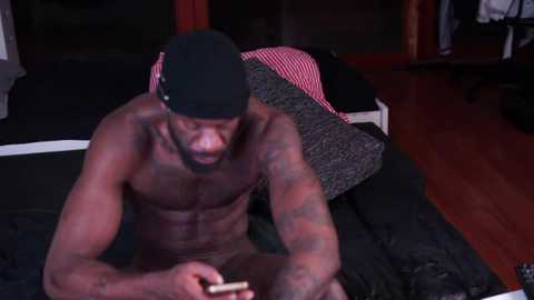 Media: Video of a shirtless, muscular, dark-skinned Black man with tattoos, wearing a black beanie, sitting on a bed with black and red striped sheets, holding a cigarette.