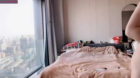 Media: A video of a modern, well-lit bedroom with a large window revealing a cityscape. The room features a bed with beige sheets, a vanity table with a mirror, and a chair with red cushions.