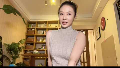 Media: Video of an East Asian woman with fair skin, black hair in a bun, wearing a sleeveless, light grey turtleneck dress, standing in a warmly lit, bookshelf-filled room with a fern and framed photos on the walls.