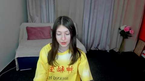 Media: Video of a young Asian woman with long dark hair, wearing a yellow shirt, sitting in a dimly lit room with white curtains and a bed with pink and blue bedding.