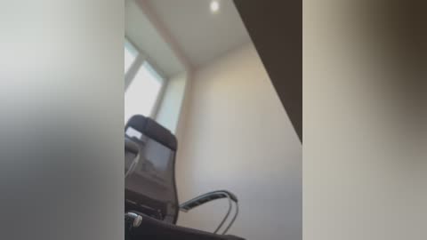 Media: A video of a minimalist office space featuring a black mesh office chair with a black metal frame, positioned in front of a large window with light streaming in. The walls are painted in a light beige color, and the ceiling is bright with recessed lighting.