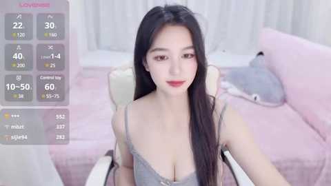 Video of an East Asian woman with long black hair, wearing a gray lace bralette, sitting on a pastel pink couch, indoors with soft lighting and a digital temperature display.