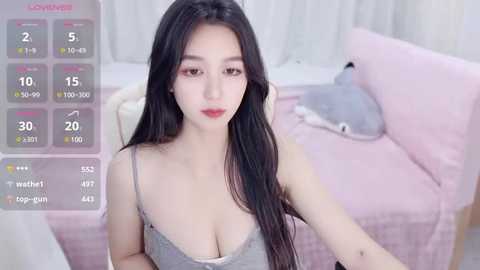 Media: Video of an East Asian woman with long black hair, light skin, and full lips, wearing a low-cut gray tank top, sitting on a pink bed. The background includes a gray teddy bear and a white chair.