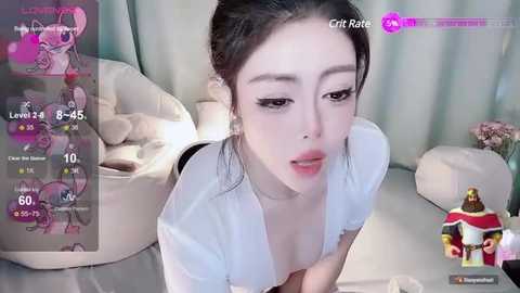 Media: A video of an Asian woman with fair skin and dark hair, wearing a white blouse, leaning forward in a cozy room with a pink stuffed animal and a plush chair.