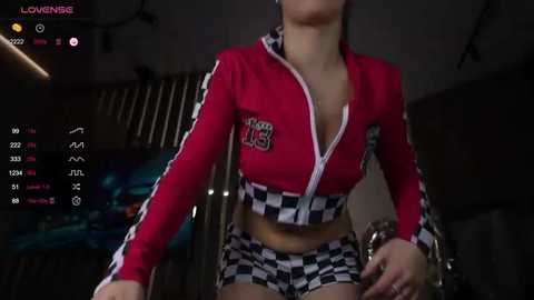 Media: Video of a young woman in a red, checkered zip-up crop top and matching shorts, partially unzipped, indoors.