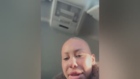 Media: Video of a young, bald, light-skinned baby with a round face, wearing a brown coat, looking up in a car with a beige ceiling and overhead air vents.