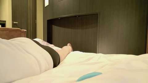 Media: Video of a person sleeping under a white bedsheet, wearing a black tank top, with a brown wooden headboard and a small blue object beside them.