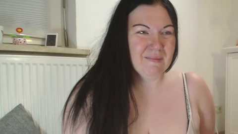 Media: Video of a smiling, pale-skinned woman with long black hair and a thin build, wearing a light-colored tank top, standing in a kitchen with white cabinets and a window with closed blinds.