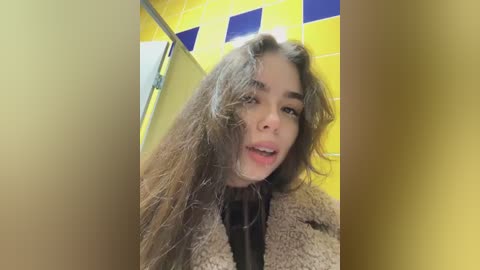 Media: A video of a young woman with long, wavy brown hair and fair skin, wearing a beige fur coat and a black top. The background features bright yellow and blue tiled walls.