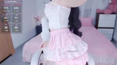 Media: Video of a slender, light-skinned woman with long, wavy black hair, wearing a white lace crop top and pink floral skirt, posing in a pink bedroom with a bed and dresser in the background.