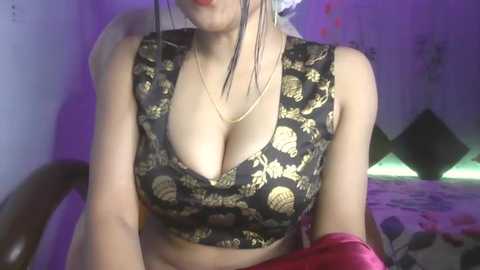 Media: Video of a woman with light skin, wearing a low-cut black floral crop top revealing ample cleavage, sitting in a dimly lit bedroom with purple lighting.