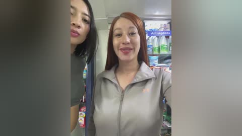 Media: Video of two women smiling, one with short black hair and the other with long red hair, both in grey uniforms, standing in a brightly lit convenience store with shelves of products in the background.