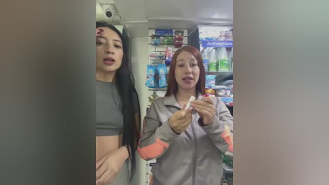 Media: Video of two women in a grocery store, one with long black hair in a grey crop top, the other with red hair in a grey jacket, both holding a box of condoms.