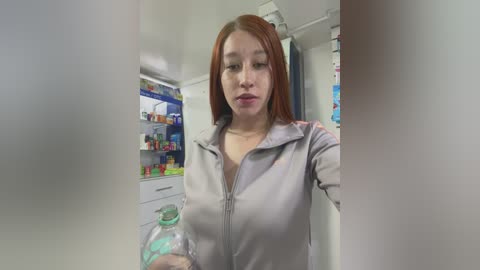 Media: A video of a young Asian woman with straight red hair, wearing a light gray zip-up hoodie, holding a clear plastic water bottle. The background shows a well-lit pharmacy with shelves of colorful medicines.