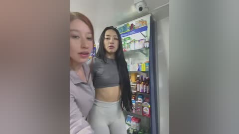 Media: Video of two women, one in a grey crop top and white pants, standing in a brightly lit convenience store. The other woman is blurred, partially visible in the foreground.