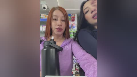 Media: Video of two young women, one with red hair and light skin, and another with long black hair, smiling and posing in a convenience store with a colorful background of snacks and drinks.