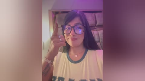 Media: A video of a smiling, light-skinned Latina woman with long dark hair, wearing glasses and a white T-shirt, standing indoors with a cozy background of beige pillows.