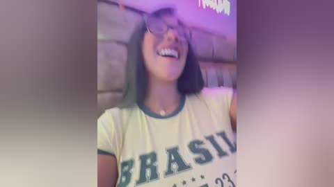 Media: Video of a young woman with straight, dark hair, wearing a yellow \"BRAZIL\" t-shirt, smiling with glasses, in a dimly lit, brick-walled room, holding a smartphone.