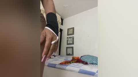 Media: A video showing a dark-skinned person's hand reaching over a bed with a blue and white checkered blanket, surrounded by framed photos and a colorful, patterned cushion on the wall.