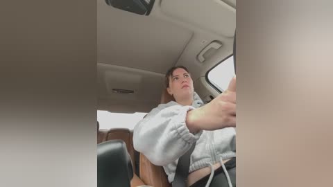 Media: A video of a young woman with light skin, brown hair, and a light grey hoodie driving a car, seen through the windshield. The interior is beige with brown leather seats.