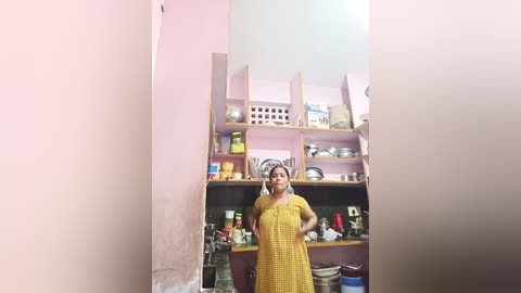 Media: Video of a middle-aged woman wearing a yellow checkered dress, standing in a small, cluttered kitchen with open shelves holding various utensils and spices.