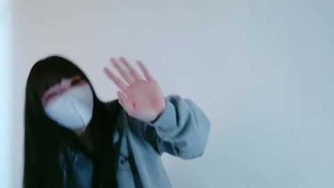 Media: Video of an Asian woman with long black hair, wearing a white mask and blue jacket, waving her hand against a plain white background.