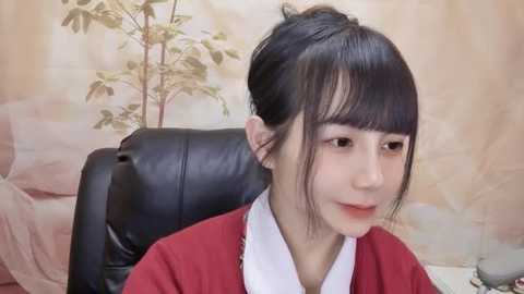 Media: A video of a young Asian woman with fair skin, black hair in a bun, wearing a red kimono, sitting on a black leather chair. Background features a floral wallpaper and a green plant.