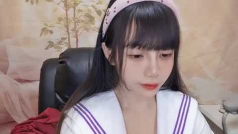 Media: Video of an Asian woman with long black hair and a white headband, wearing a Japanese schoolgirl uniform. She sits on a black leather chair in a room with beige walls and a floral wallpaper.