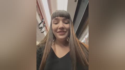 Media: Video of a smiling, light-skinned woman with long, straight brown hair styled with blunt bangs, wearing a black V-neck shirt, standing in a dimly lit hallway with white walls and a door handle.