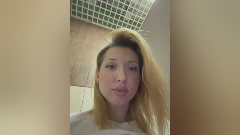 Media: A video of a blonde woman with light skin, wearing a white shirt, standing in a bathroom with a beige-tiled wall and a mosaic-tiled ceiling.